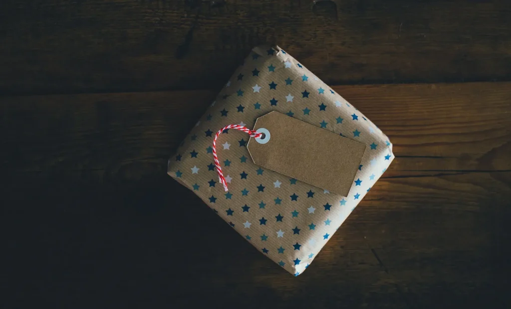 Pacco regalo avvolto in carta decorata con piccole stelle blu e bianche, appoggiato su una superficie in legno scuro. Sul pacco è fissata un’etichetta marrone vuota con cordoncino bianco e rosso.