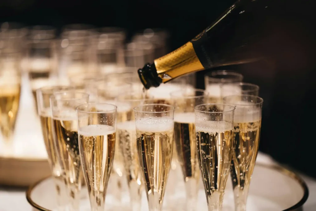 Bottiglia di spumante inclinata mentre versa il contenuto in una serie di flute disposti su un vassoio. I bicchieri sono parzialmente riempiti e mostrano bollicine e schiuma in superficie; sullo sfondo sono visibili altri calici sfocati.