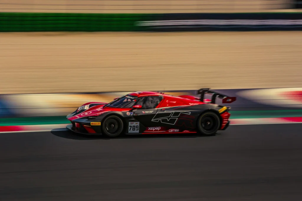 Auto da corsa rossa e nera in movimento su un circuito automobilistico. Il veicolo presenta il numero 789 sulla fiancata ed è ripreso lateralmente mentre percorre un tratto asfaltato delimitato da cordoli colorati e barriere di protezione.