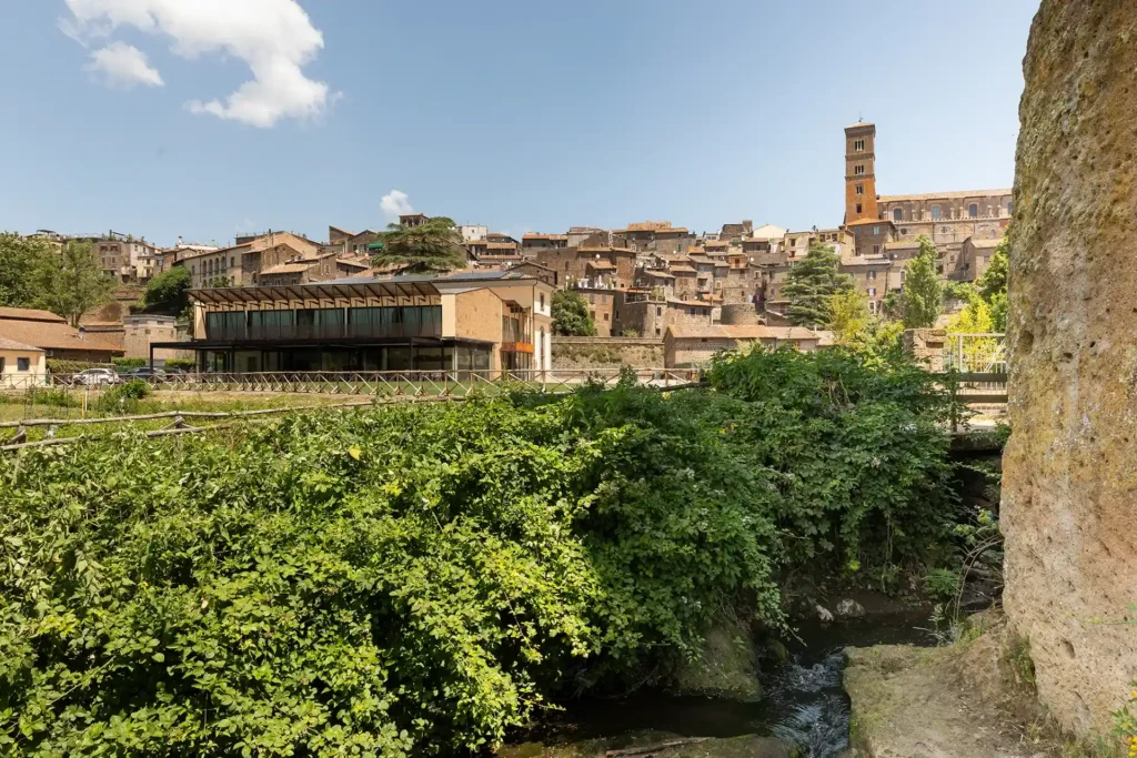Vista dell’hotel e del centro storico circostante con edifici in pietra e campanile visibile sullo sfondo. In primo piano si trovano vegetazione, un piccolo corso d’acqua e una parete rocciosa; l’edificio dell’hotel è visibile sulla sinistra.