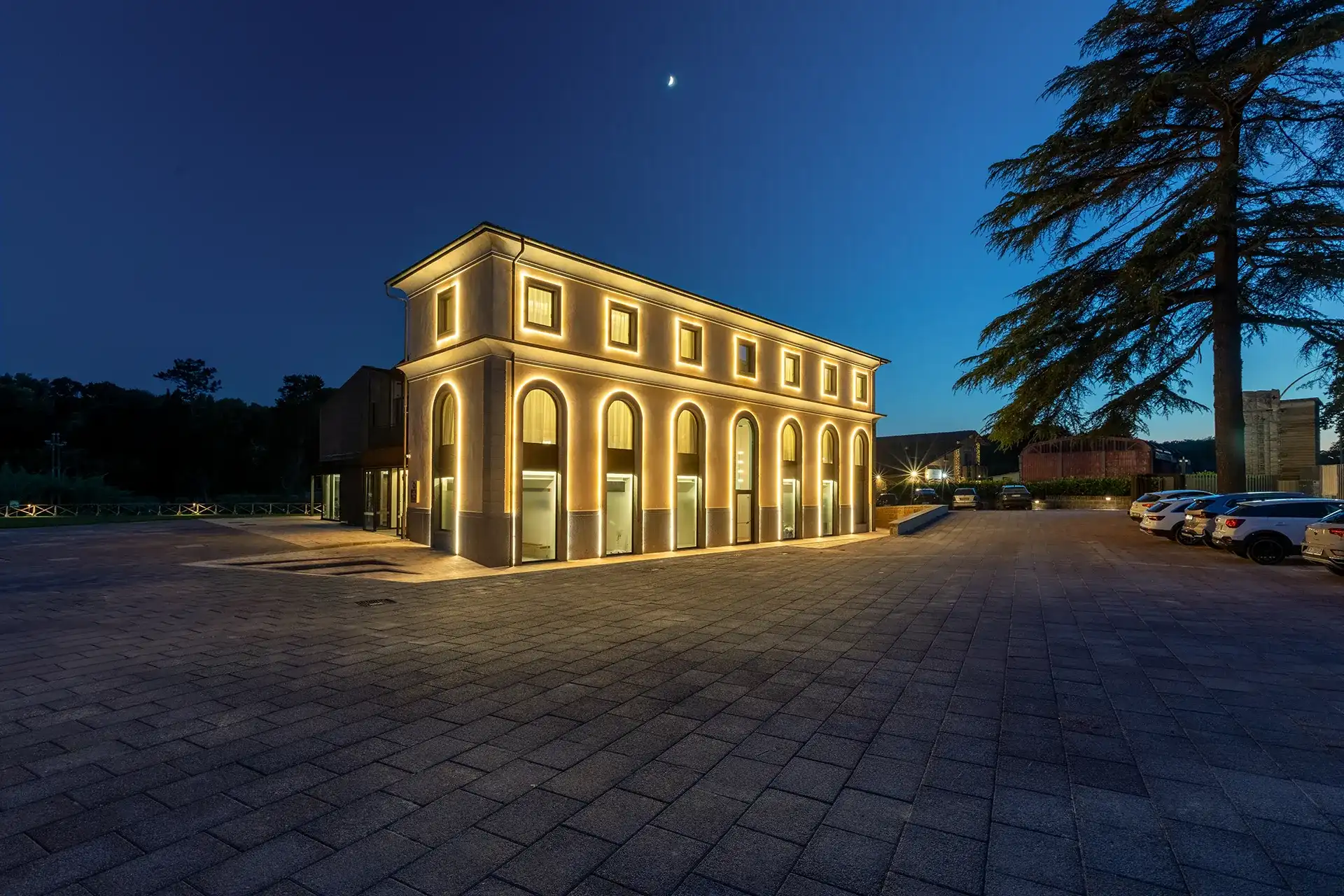 Vista esterna dell’hotel con edificio a due piani caratterizzato da grandi finestre ad arco illuminate e ampio piazzale pavimentato. A destra sono visibili alcune auto parcheggiate e un grande albero, mentre sul lato sinistro si estende l’area esterna della struttura.