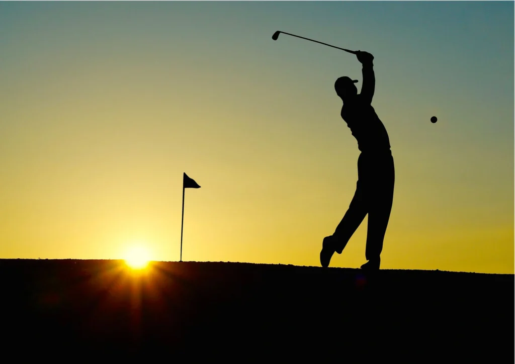 Persona ripresa in controluce mentre esegue uno swing con una mazza da golf su un campo da gioco. A sinistra è visibile una bandierina che segnala la buca, mentre il sole si trova vicino all’orizzonte e la pallina da golf è visibile in volo.