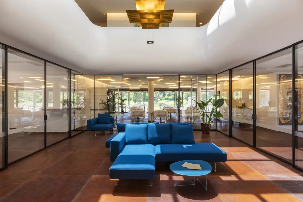 Area lounge con divano modulare blu e tavolino rotondo posizionati al centro di un ampio spazio interno. L’ambiente è delimitato da pareti vetrate che si affacciano su una sala ristorante con tavoli apparecchiati e piante in vaso. Sono visibili pavimento scuro, una lampada sospesa sul soffitto e grandi finestre sul fondo.