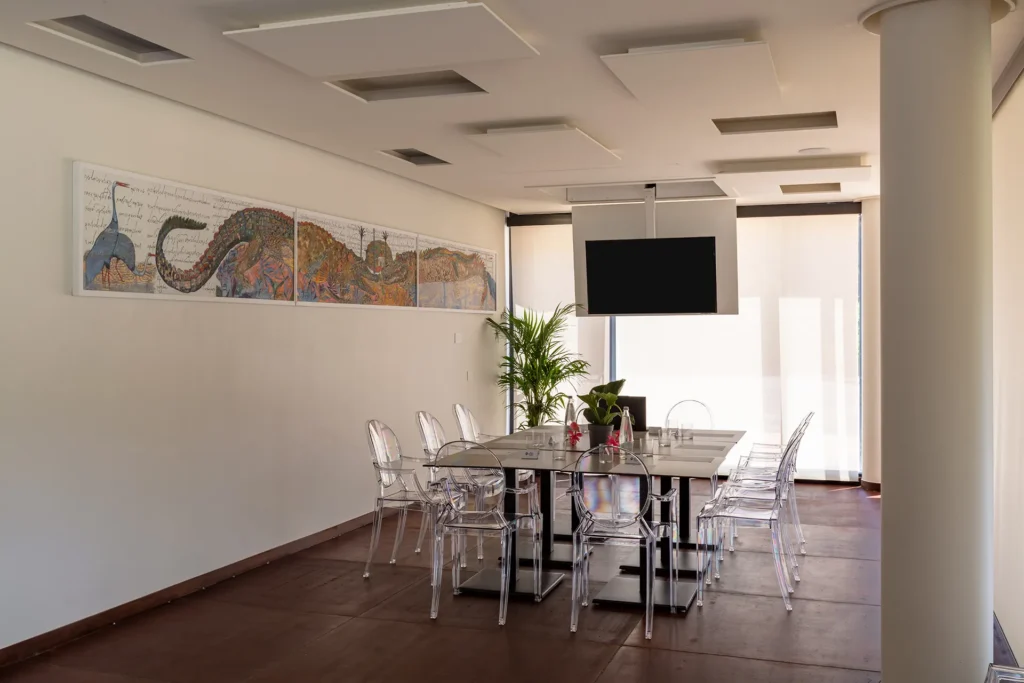 Sala meeting con file di sedie trasparenti disposte di fronte a un tavolo per relatori e a uno schermo sospeso. Sulla parete laterale è presente un lungo quadro decorativo, mentre sul fondo si vede una pianta in vaso. L’ambiente ha colonne bianche, ampie finestre con tende chiare e pavimento scuro.