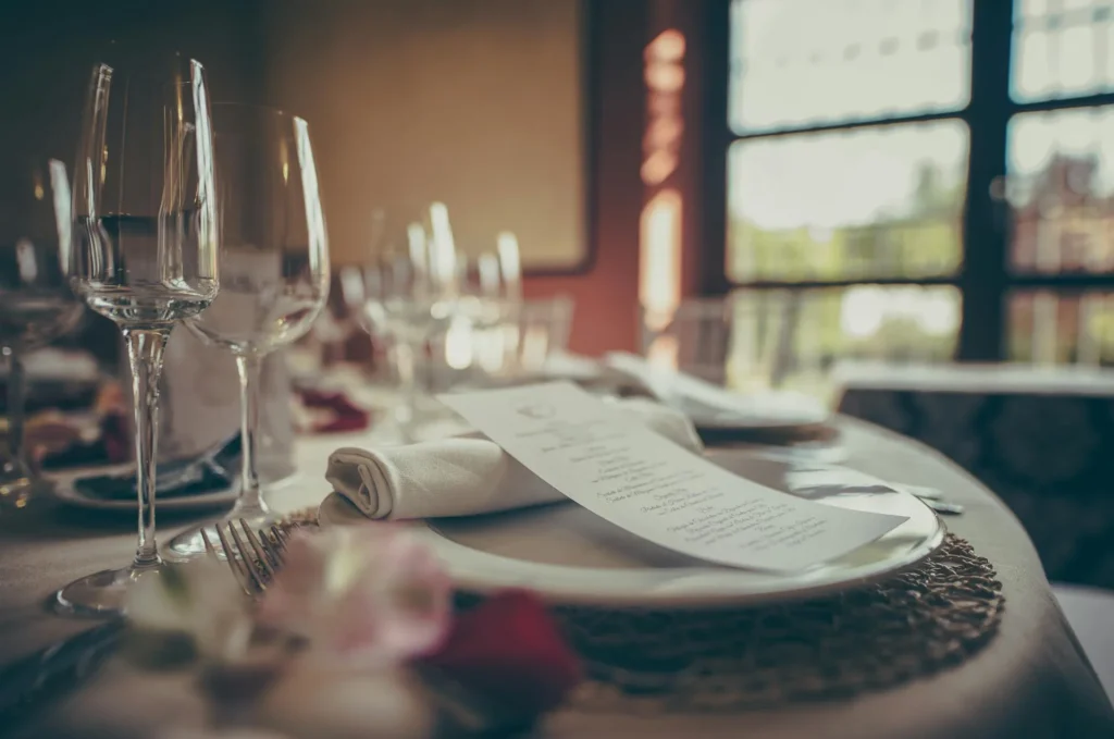 Tavolo apparecchiato con piatto, tovagliolo arrotolato, posate e calici da vino. Sul piatto è appoggiato un menu stampato, mentre sullo sfondo sono visibili altri tavoli e una grande finestra che illumina la sala ristorante.