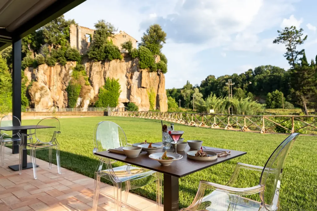 Tavolo apparecchiato su una terrazza coperta con piatti e bevande, affacciato su un ampio prato. Sullo sfondo si vede una parete rocciosa con resti di un edificio in pietra, circondata da alberi e vegetazione.