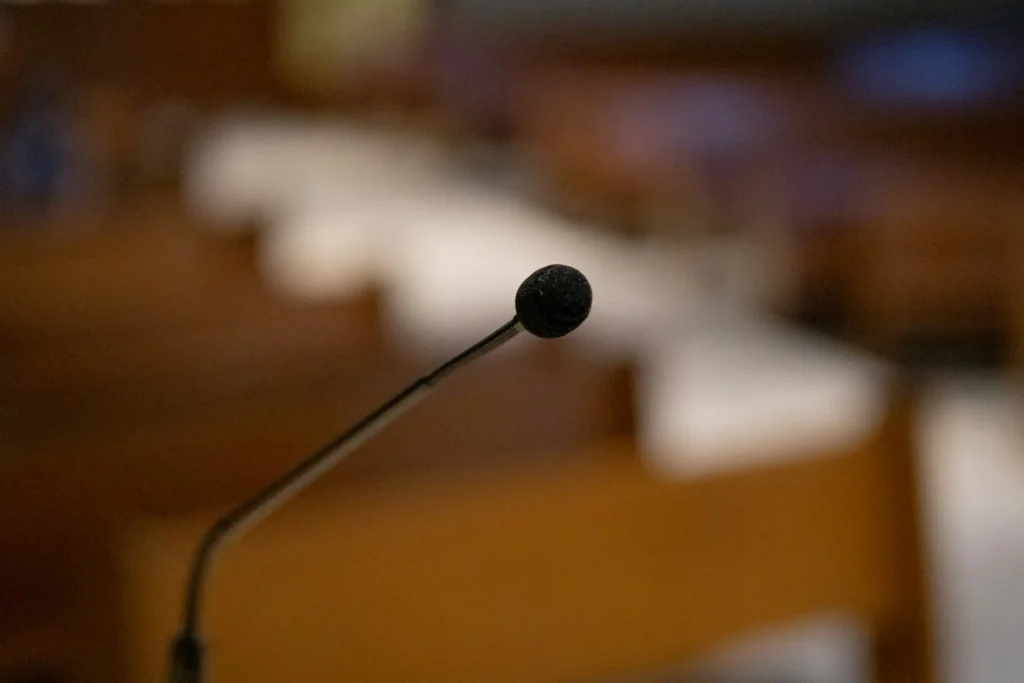 Primo piano di un microfono a collo d’oca con terminale in schiuma nera, posizionato in una sala conferenze o riunioni. Sullo sfondo, sfocati, sono visibili sedute e arredi in legno disposti in file.