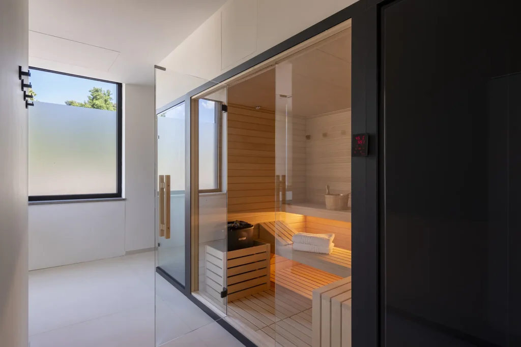 Piccola area benessere con sauna in legno dotata di pareti e porta in vetro. All'interno della sauna si vedono panche in legno, asciugamani piegati e accessori per il bagno di calore. La stanza ha pareti chiare, pavimento chiaro e una grande finestra laterale con vetro opaco.