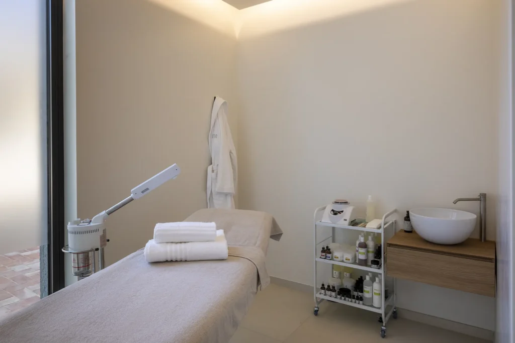 Stanza per trattamenti con lettino coperto da un telo chiaro e asciugamani piegati sopra. Accanto al lettino si trovano un carrello con prodotti e attrezzature, mentre sulla parete è appeso un accappatoio. Nella stanza sono presenti anche un lavabo con mobile in legno e una grande finestra laterale.