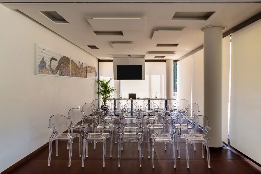 Sala conferenze con sedie trasparenti disposte in file davanti a un tavolo e a uno schermo sospeso. Lungo la parete sinistra è appeso un quadro orizzontale, mentre sul fondo si trova una pianta in vaso. La stanza presenta grandi finestre con tende chiare, colonne bianche e pavimento scuro.