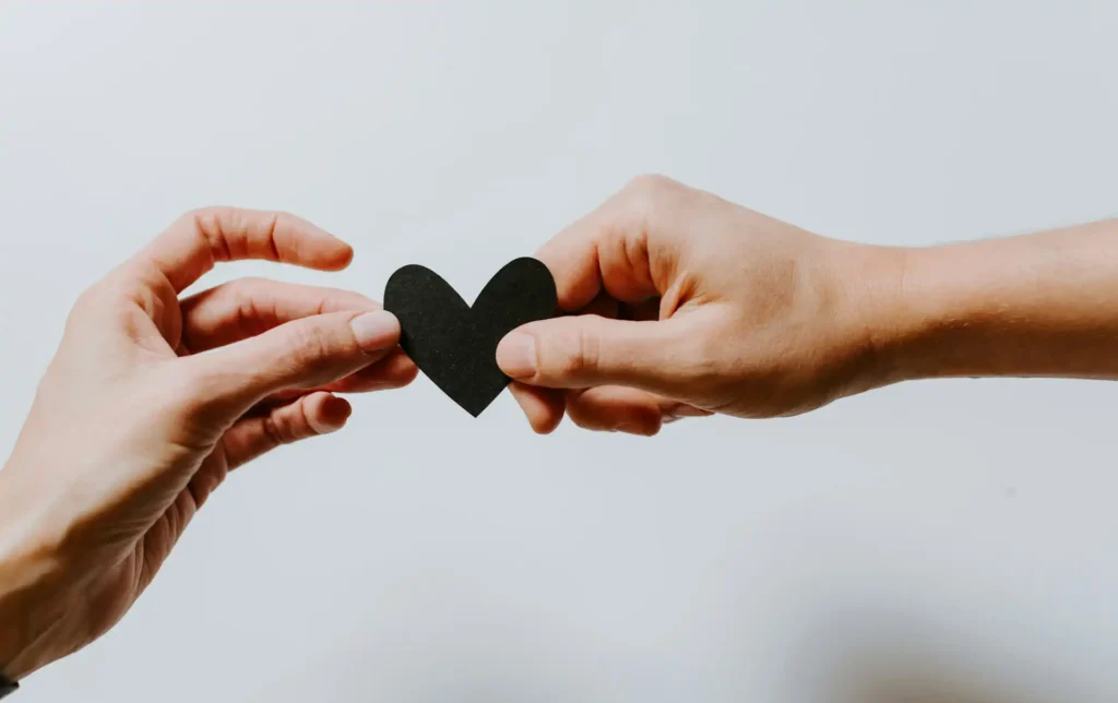 Il primo piano sulla mano di una donna e la mano di un uomo che, insieme, reggono un cuore di carta di colore nero.